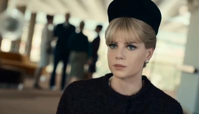 A closeup of Lucy Boynton in a 1960s style black hat and black coat, with a group of people also dressed in dark clothes out of focus in the background.